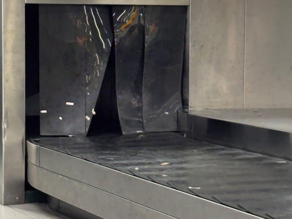 Ein leeres Gepäckband am Flughafen von Curaçao kommt aus einer Öffnung mit schwarzen Gummilamellen hervor. Die Szene zeigt den metallischen Rahmen und die dunkle Oberfläche des Förderbands in einer sachlichen Flughafenatmosphäre.