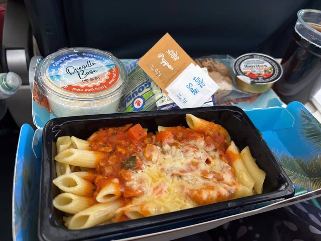 Ein Tablett mit Flugzeugessen auf einem Flug nach Curaçao, bestehend aus Penne-Nudeln mit Tomatensoße und geschmolzenem Käse, einem Dessertbecher „Quesillo Base“, Käse, Salz, Pfeffer und einem Getränk.