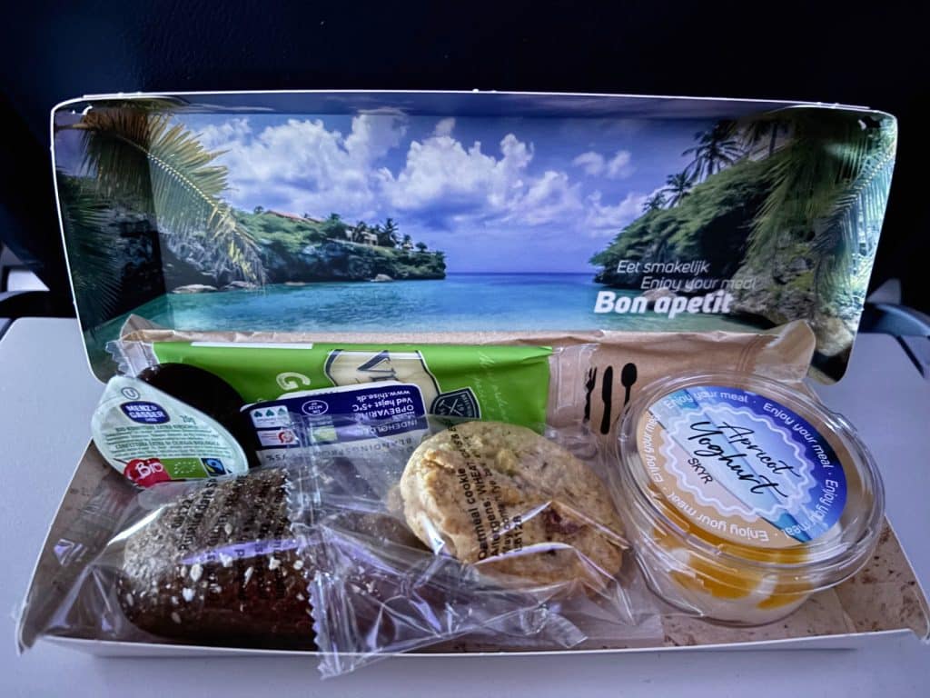 Ein Frühstückspaket auf einem Flug nach Curaçao in einem Pappkarton mit tropischem Strandmotiv, bestehend aus einem dunklen Brötchen, einem Haferkeks, Aprikosen-Skyr, Marmelade und Besteck.