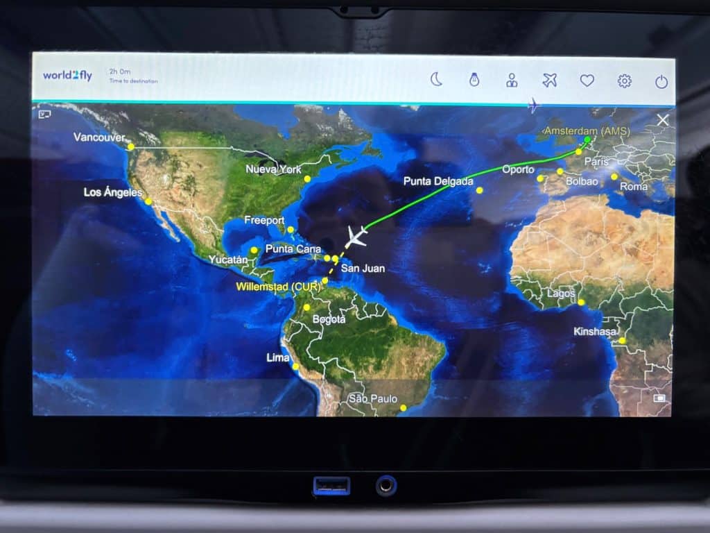 Ein Bordmonitor in einem Flugzeug zeigt eine Weltkarte mit der Flugroute von Amsterdam nach Curaçao, wobei sich das Flugzeugsymbol aktuell über dem Atlantik befindet. Auf der Karte sind verschiedene Städte markiert, darunter Punta Cana, San Juan und Willemstad (CUR).