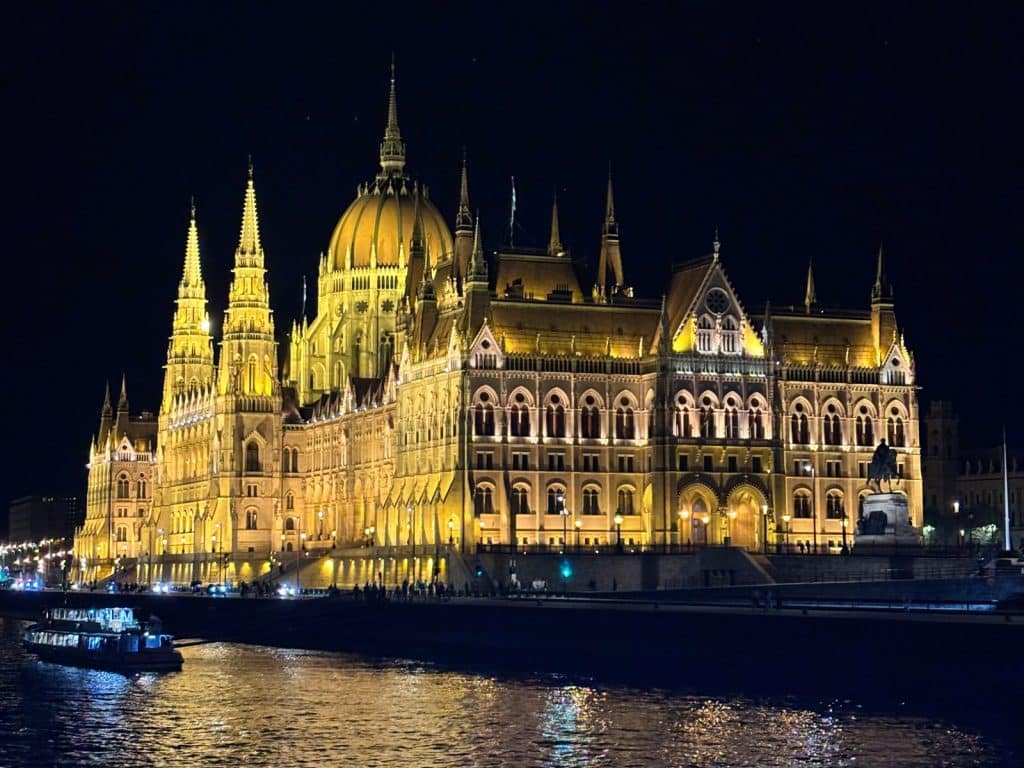 Das beleuchtete ungarische Parlamentsgebäude in Budapest erstrahlt nachts in goldenem Licht am Ufer der Donau, während im Vordergrund ein hell erleuchtetes Flusskreuzfahrtschiff vorbeifährt.