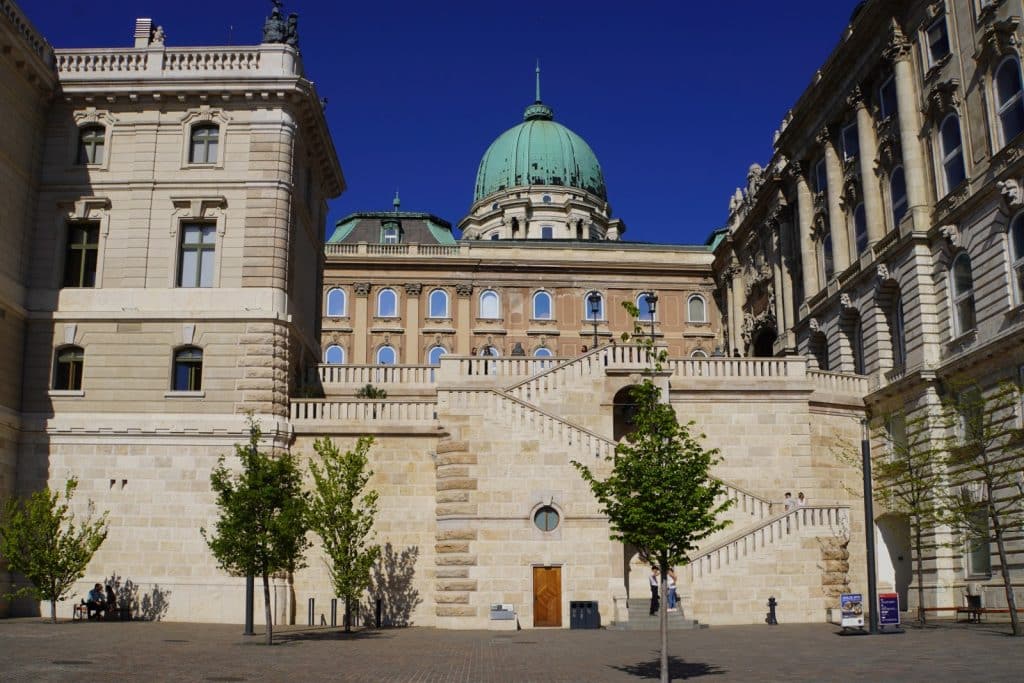 Blick auf den gepflasterten Innenhof der Burg in Budapest mit einer monumentalen Freitreppe aus hellem Stein, die vor der markanten grünen Kuppel des Schlosses emporragt.