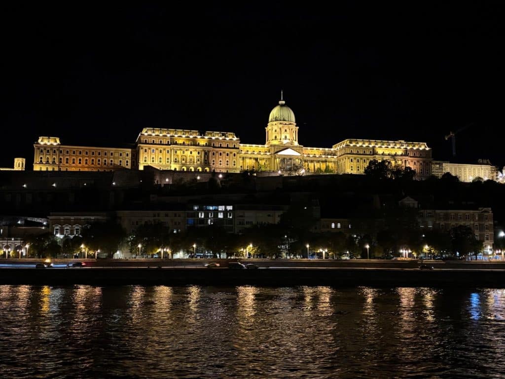 Eine nächtliche Ansicht der beleuchteten Budapester Burg, die auf einem Hügel über der Donau thront und sich golden im dunklen Wasser spiegelt. Am Ufer unterhalb der Burgmauer sind Lichter einer Uferstraße und parkende Autos vor dunklen Gebäudefassaden zu erkennen.
