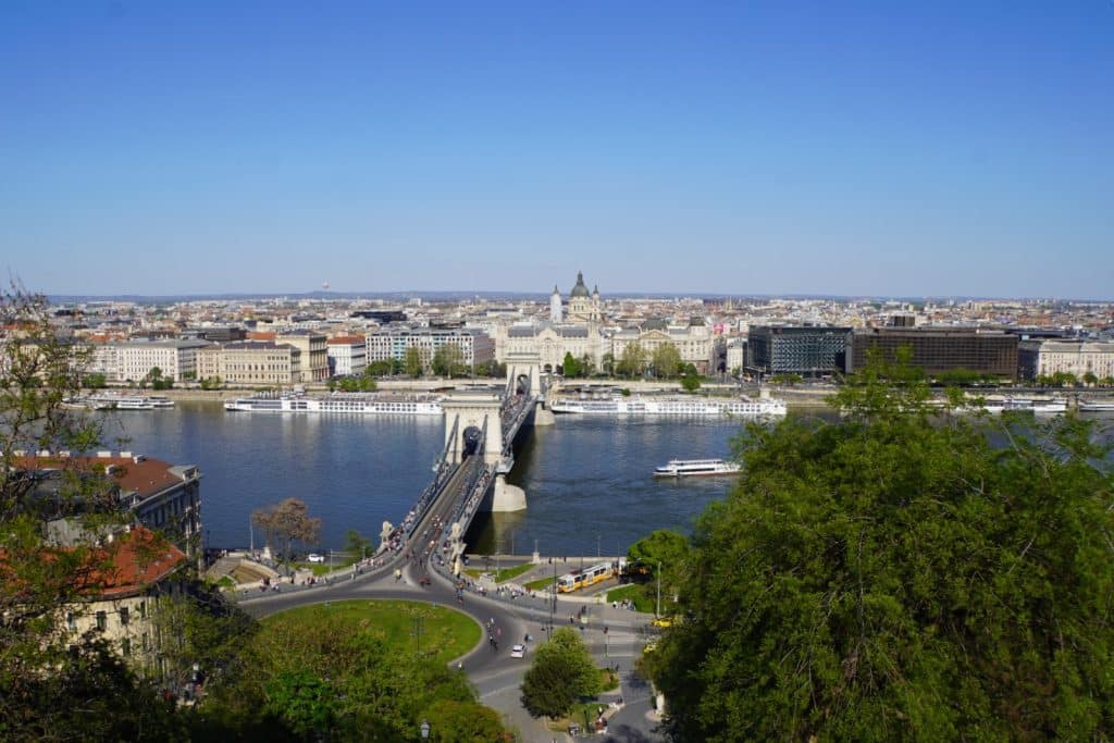 Blick von einer Anhöhe auf die Donau in Budapest mit der Széchenyi-Kettenbrücke, mehreren Flusskreuzfahrtschiffen am Ufer und der St.-Stephans-Basilika im Hintergrund.