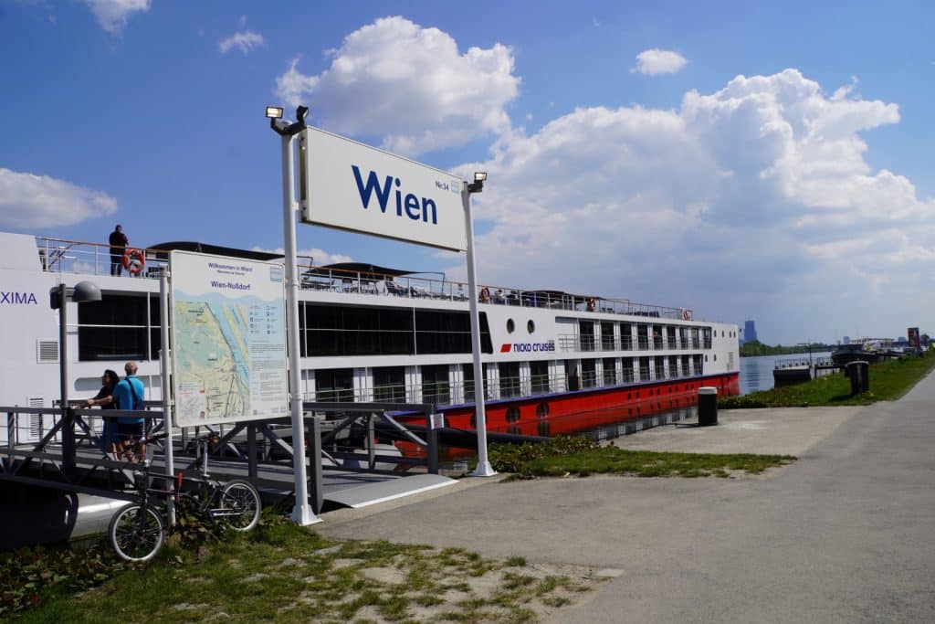 Ein großes, weiß-rotes Flusskreuzfahrtschiff der Reederei nicko cruises liegt bei sonnigem Wetter an der Anlegestelle Wien-Nußdorf an der Donau vor Anker. Im Vordergrund sind ein Hinweisschild mit der Aufschrift „Wien“, ein Stadtplan sowie Fahrräder und Passagiere auf dem Steg zu sehen.