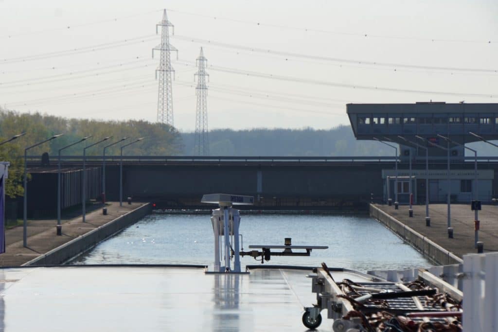 Blick vom Deck eines Flusskreuzfahrtschiffes auf der Donau in eine Schleusenkammer vor Wien, mit Kontrollgebäuden am Ufer und Hochspannungsmasten im Hintergrund.