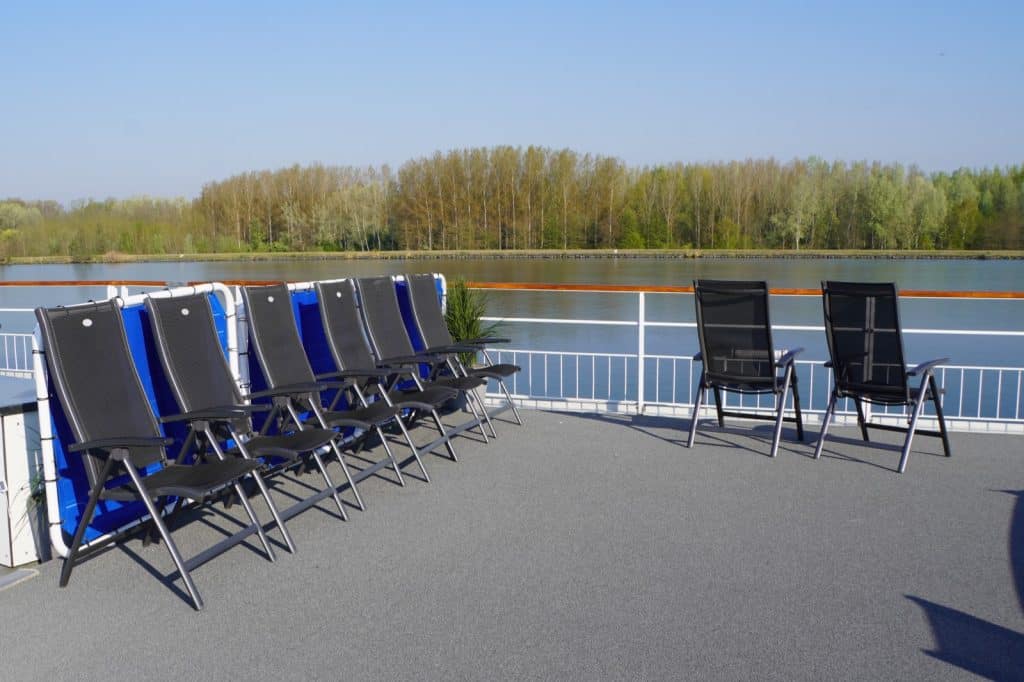 Auf dem Sonnendeck eines Flusskreuzfahrtschiffes auf der Donau stehen mehrere dunkle Klappstühle vor einer weißen Reling mit Blick auf den Fluss und ein bewaldetes Ufer.