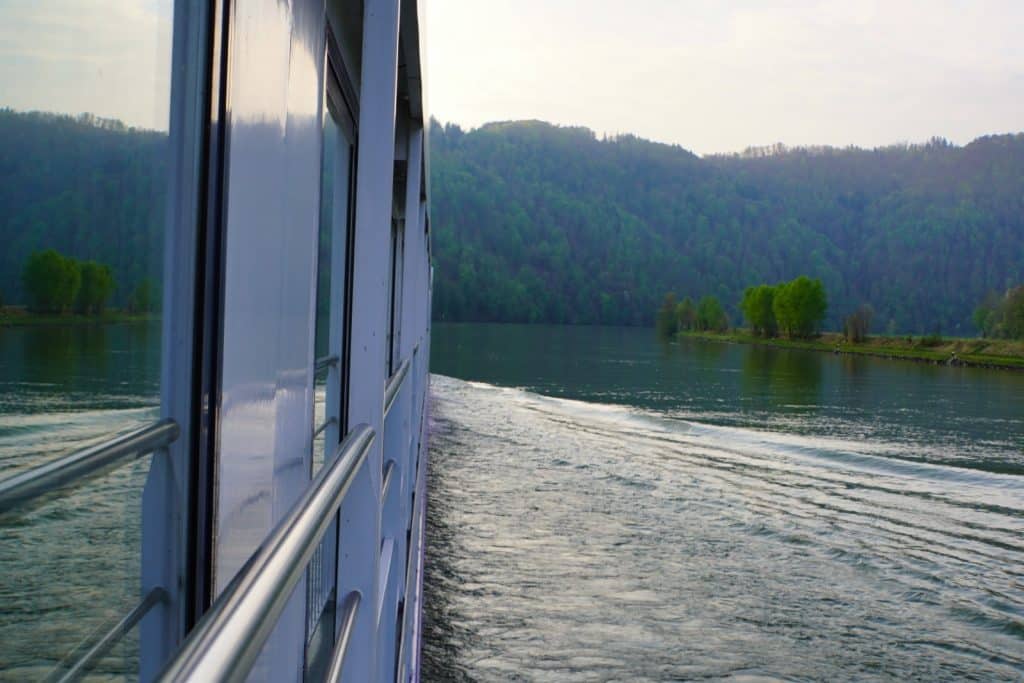 Blick von der Reling eines Kreuzfahrtschiffes auf die Donau bei Passau am Abend, während das Schiff eine sanfte Heckwelle im ruhigen Wasser vor einer bewaldeten Hügelkette hinterlässt.
