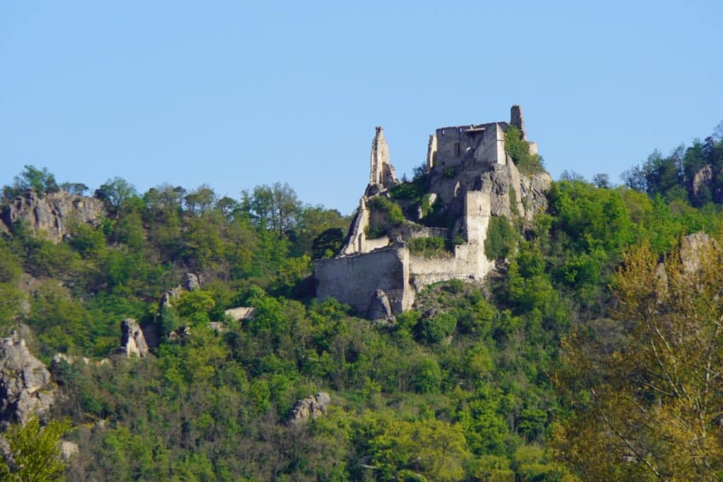 Die Ruine Dürnstein thront auf einem bewaldeten Felshügel über der Donau in der Nähe von Melk, während ihre verfallenen Steinmauern und Türme in den blauen Himmel ragen.