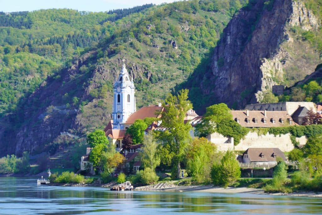 Blick von einer Flusskreuzfahrt auf der Donau auf das Stift Dürnstein mit seiner markanten blauen Kirche vor einer bewaldeten Hügellandschaft.