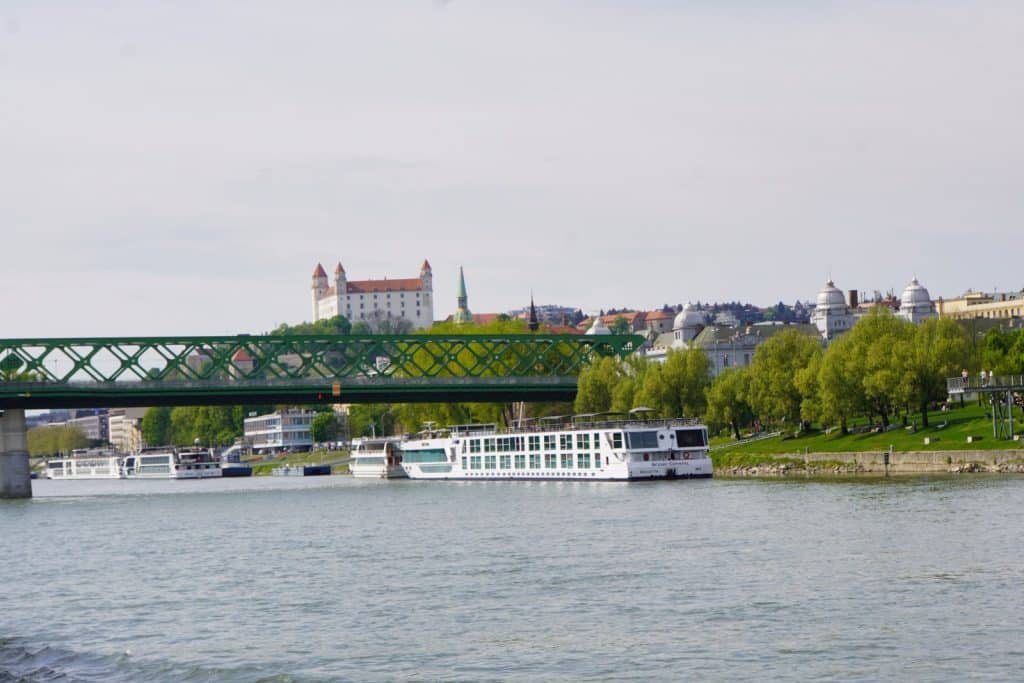 Ein großes weißes Flusskreuzfahrtschiff fährt auf der Donau in Bratislava ein, im Hintergrund sind die markante Burg Bratislava auf einem Hügel und die grüne Alte Brücke zu sehen.