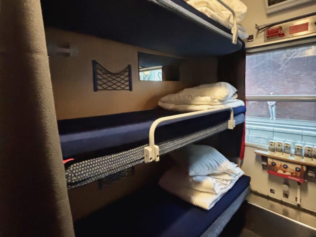 Innenansicht eines Snälltåget-Reisezug Abteils mit drei übereinander angeordneten Schlafkojen, die mit blauen Matratzen und weißem Bettzeug ausgestattet sind. Neben den Betten befindet sich ein Fenster mit Blick auf einen Bahnsteig sowie eine kleine Ablage mit Getränkekartons.