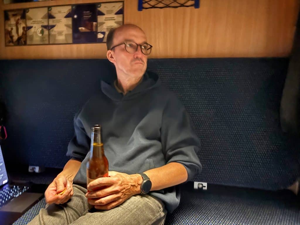 Ein Mann mit Brille und Kapuzenpullover sitzt in einem Zugabteil im Snälltåget-Reisezug, hält eine geöffnete Bierflasche in der Hand und blickt nachdenklich zur Seite.
