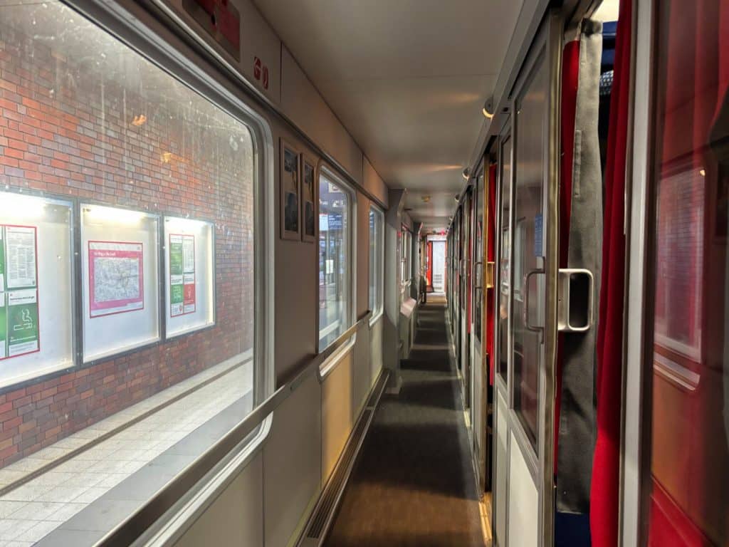 Blick durch den schmalen Gang eines Snälltåget-Reisezug, mit großen Fenstern auf der linken Seite, die eine Ziegelwand am Bahnsteig zeigen, und Abteiltüren mit roten Vorhängen auf der rechten Seite.
