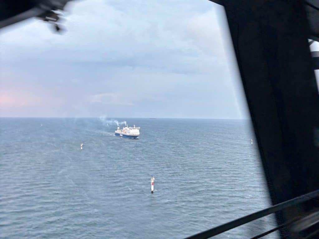 Blick aus einem Zug auf der Öresundbrücke auf eine blau-weiße Fähre, die mit rauchenden Schornsteinen auf dem weiten Meer fährt.