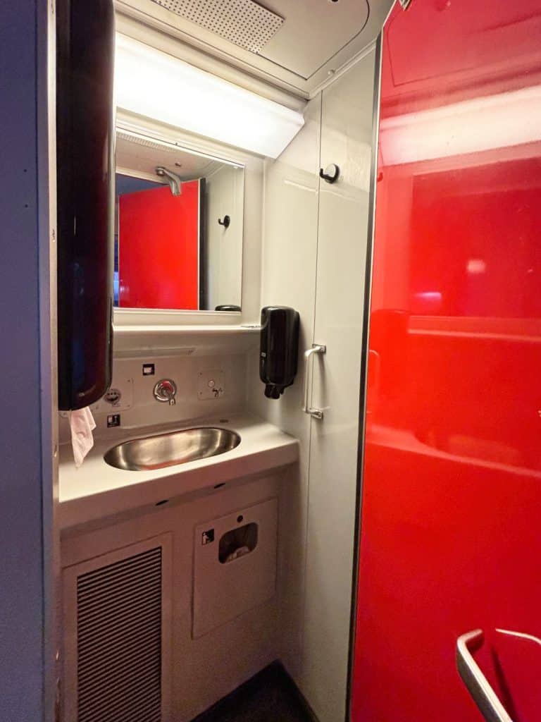 Ein kompakter Waschraum in einem Snälltåget-Reisezug mit einem silbernen Waschbecken, einem Spiegel und einer markanten roten Tür. Die Kabine ist funktional mit Spendern für Seife und Papierhandtücher sowie einer hellen Deckenbeleuchtung ausgestattet.