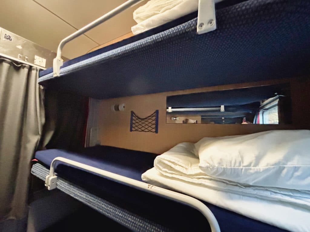 Innenansicht eines Schlafwagenabteils im Snälltåget-Reisezug mit zwei übereinander angeordneten blauen Etagenbetten, die mit weißem Bettzeug ausgestattet sind.