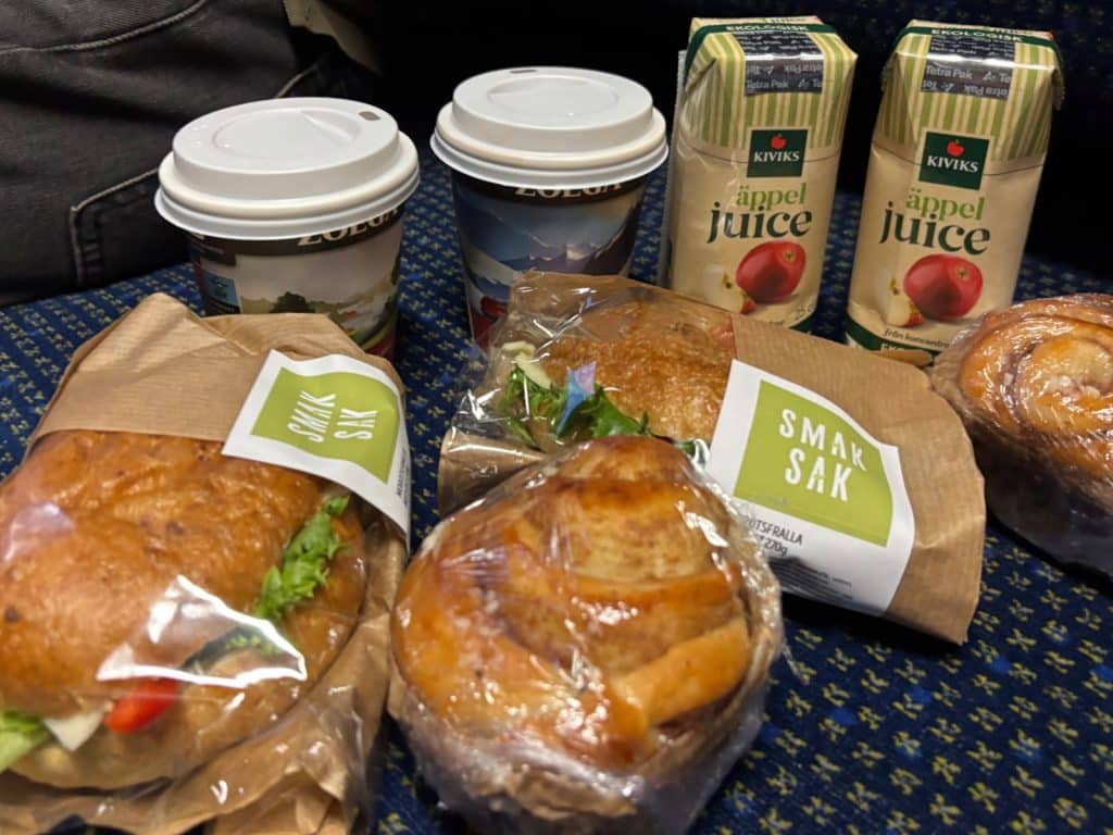 Ein Frühstück im Zug nach Malmö, bestehend aus zwei belegten Brötchen in Papiertüten der Marke Smak Sak, zwei Zimtschnecken, zwei Bechern Kaffee und zwei Packungen Kiviks Apfelsaft auf einem blauen Polstersitz.