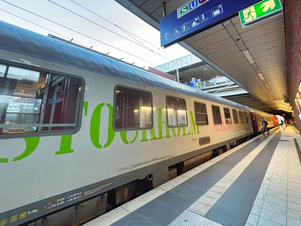 Ein weißer Reisezug der Snälltåget mit der grünen Aufschrift „STOCKHOLM“ und der rosa Aufschrift „MALMÖ“ steht an einem Bahnsteig in Berlin. Über dem Zug hängt ein blaues Bahnhofsschild mit Piktogrammen und einem grünen Notausgangsschild.