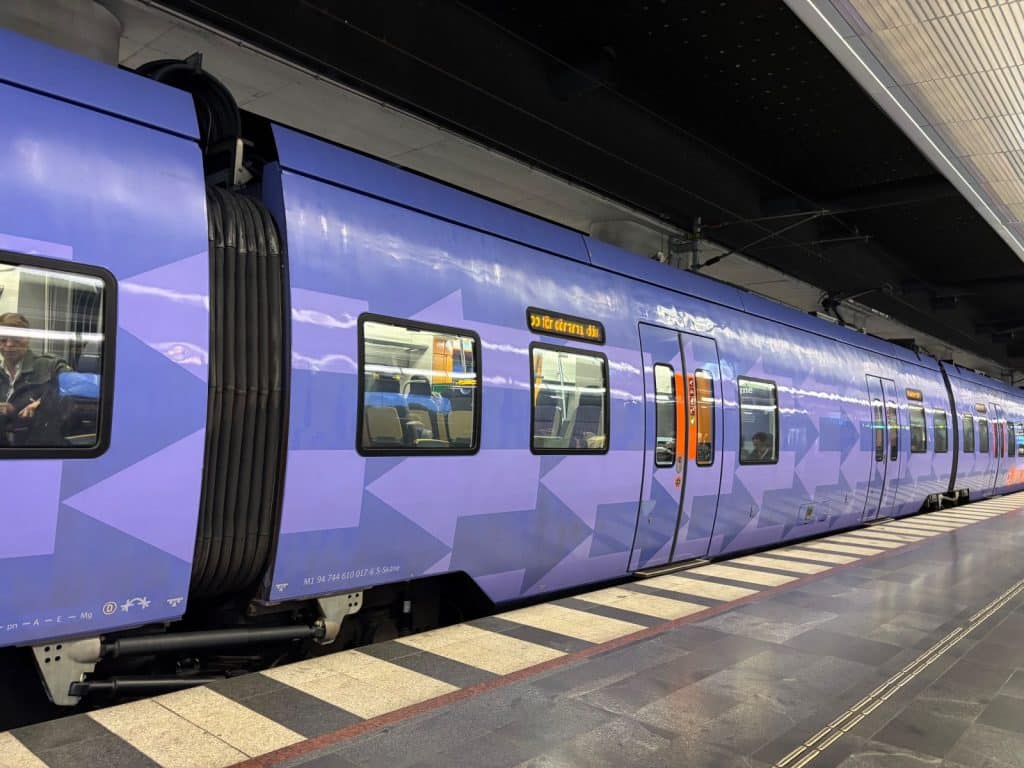 Ein lila Regionalzug nach Landskrona steht an einem modernen Bahnsteig unter einer dunklen Überdachung. Die Waggons sind mit einem geometrischen Pfeilmuster verziert und zeigen beleuchtete Fenster sowie eine digitale Zielanzeige.