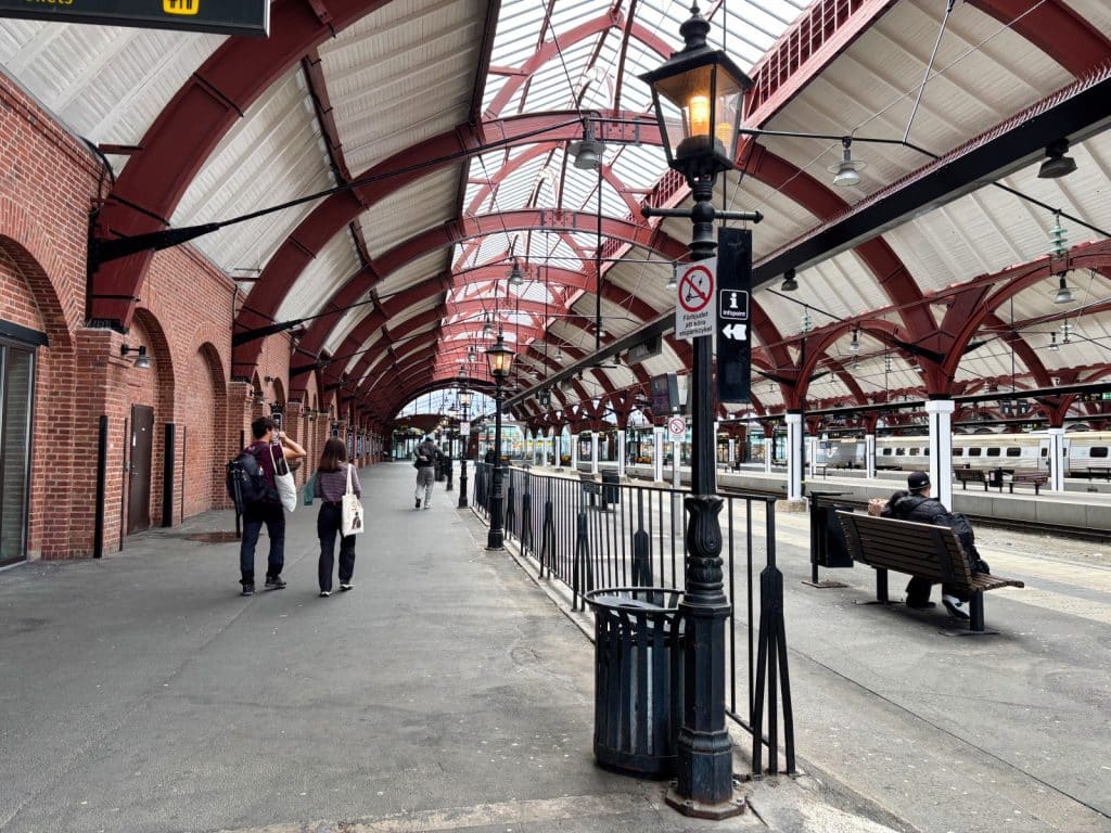 Ein Blick entlang eines Bahnsteigs im Bahnhof von Malmö unter einer hohen, bogenförmigen Dachkonstruktion aus roten Stahlträgern, auf dem Reisende spazieren und eine Person auf einer Holzbank sitzt.