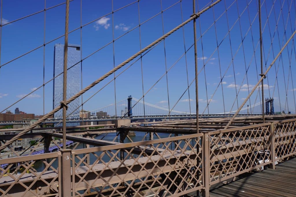 Blick von der hölzernen Promenade der Brooklyn Bridge durch die markanten Stahlseile und Geländer auf die Manhattan Bridge und einen modernen Wolkenkratzer unter blauem Himmel.