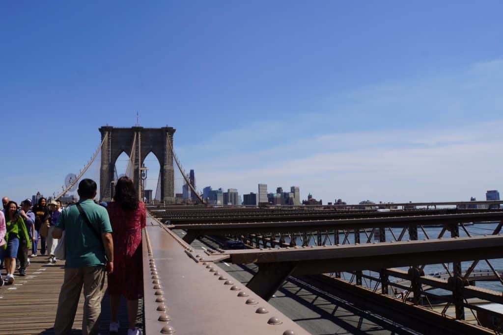 Blick von der hölzernen Fußgängerpromenade der Brooklyn Bridge auf die stählerne Fahrbahnkonstruktion und einen der steinernen Brückenpfeiler unter strahlend blauem Himmel.