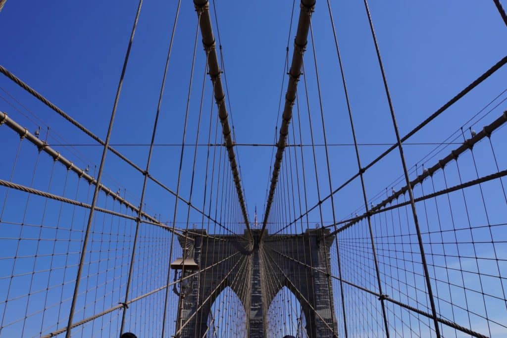 Blick von unten auf die symmetrisch verlaufenden Seile und Kabel der Brooklyn Bridge vor einem klaren, blauen Himmel, die auf einen der steinernen Brückenpfeiler zulaufen.