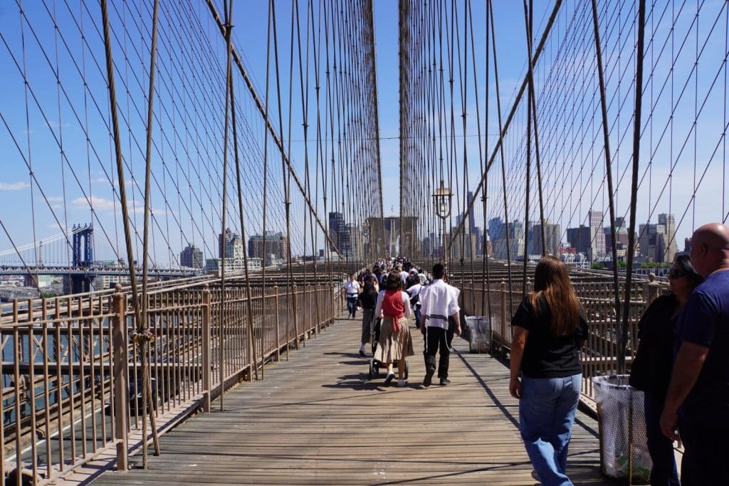 Zahlreiche Menschen spazieren über den hölzernen Fußgängerweg der Brooklyn Bridge, die von markanten Stahlseilen und hohen Geländern gesäumt ist, während im Hintergrund die Skyline von New York City und eine weitere Brücke unter blauem Himmel zu sehen sind.