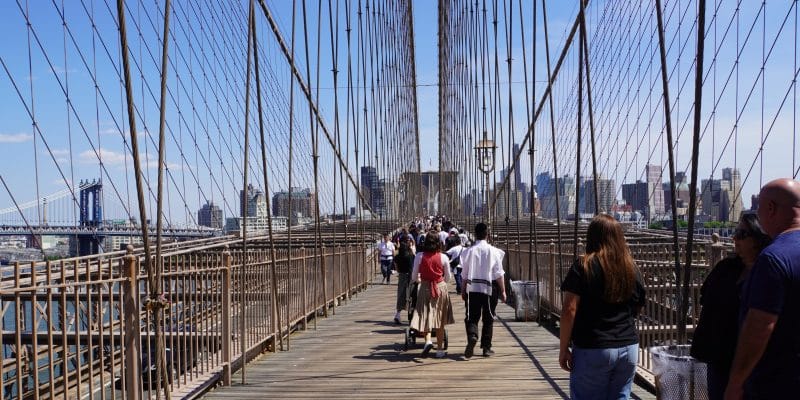 Zahlreiche Menschen spazieren über den hölzernen Fußgängerweg der Brooklyn Bridge, die von markanten Stahlseilen und hohen Geländern gesäumt ist, während im Hintergrund die Skyline von New York City und eine weitere Brücke unter blauem Himmel zu sehen sind.