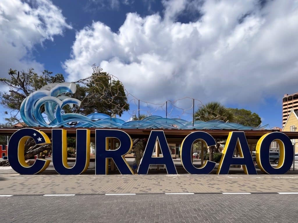 Ein großes, blau-gelbes Schild mit der Aufschrift „CURACAO“ steht auf einem gepflasterten Platz vor einer dekorativen Wellenskulptur unter einem leicht bewölkten Himmel.