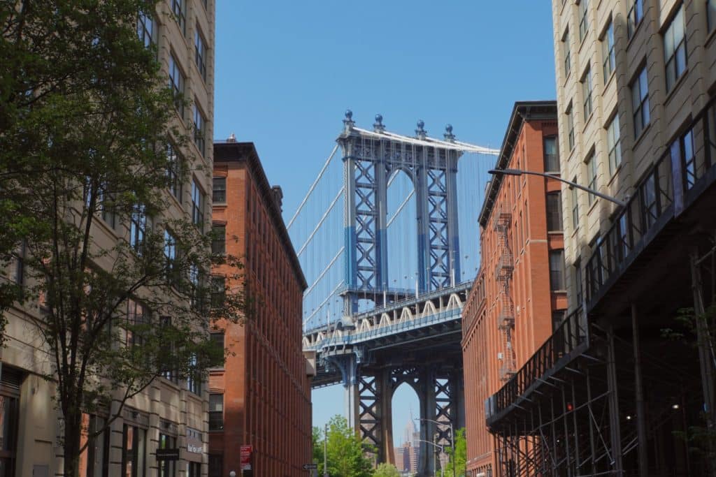 Blick aus einer Straße im New Yorker Stadtteil DUMBO, flankiert von Backsteingebäuden, direkt auf einen der blauen Stützpfeiler der Manhattan Bridge unter klarem blauem Himmel.