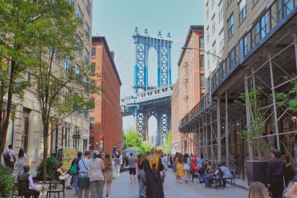 Blick durch eine belebte Straße im Viertel DUMBO in Brooklyn auf einen blauen Pfeiler der Manhattan Bridge, die von mehrstöckigen Backsteingebäuden eingerahmt wird.