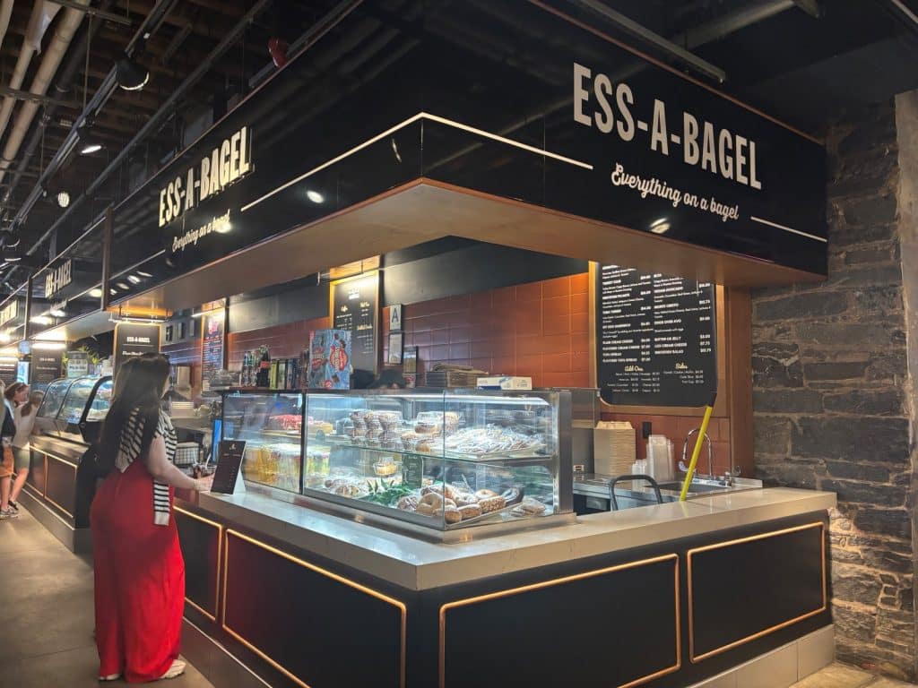 Ein Stand von Ess-A-Bagel im Time Out Market in DUMBO, Brooklyn, mit einer Auswahl an Backwaren in einer Glasvitrine und einer Kundin in einem roten Kleid davor.