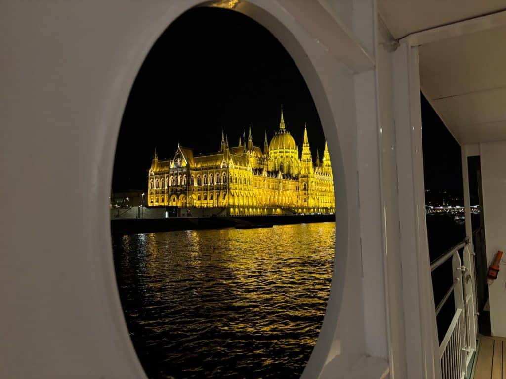 Blick durch ein ovales Schiffsfenster auf das hell erleuchtete ungarische Parlamentsgebäude in Budapest bei Nacht, dessen goldene Lichter sich im dunklen Wasser der Donau spiegeln.