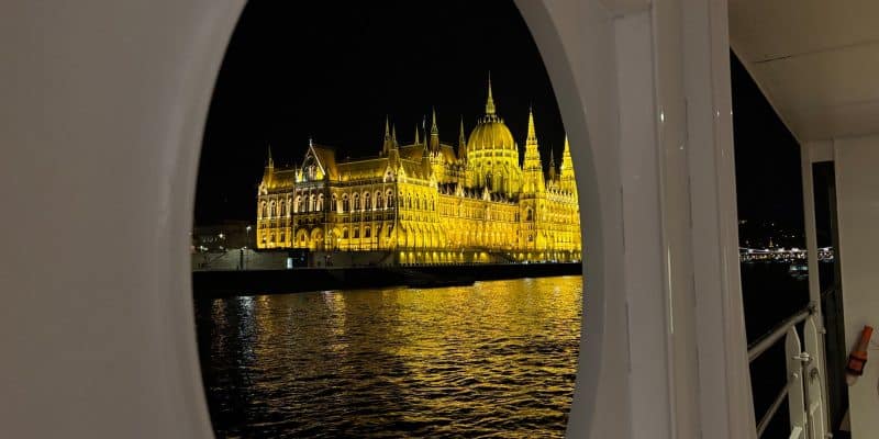 Blick durch ein ovales Schiffsfenster auf das hell erleuchtete ungarische Parlamentsgebäude in Budapest bei Nacht, dessen goldene Lichter sich im dunklen Wasser der Donau spiegeln.