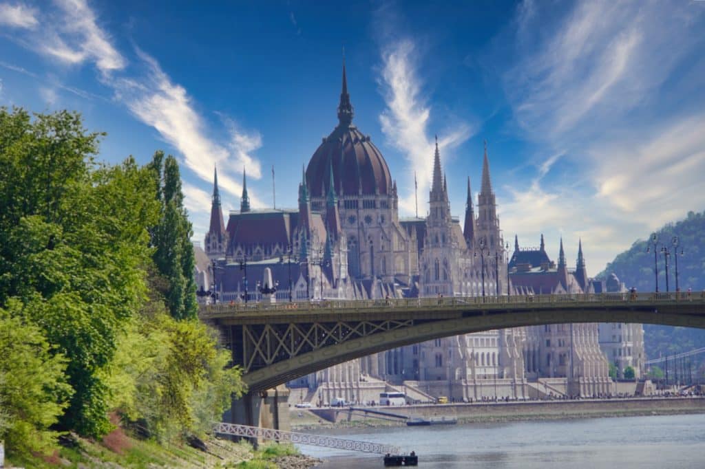 Blick vom Flussufer auf das imposante ungarische Parlamentsgebäude in Budapest, das hinter einer Brücke unter einem strahlend blauen Himmel mit Schleierwolken aufragt.