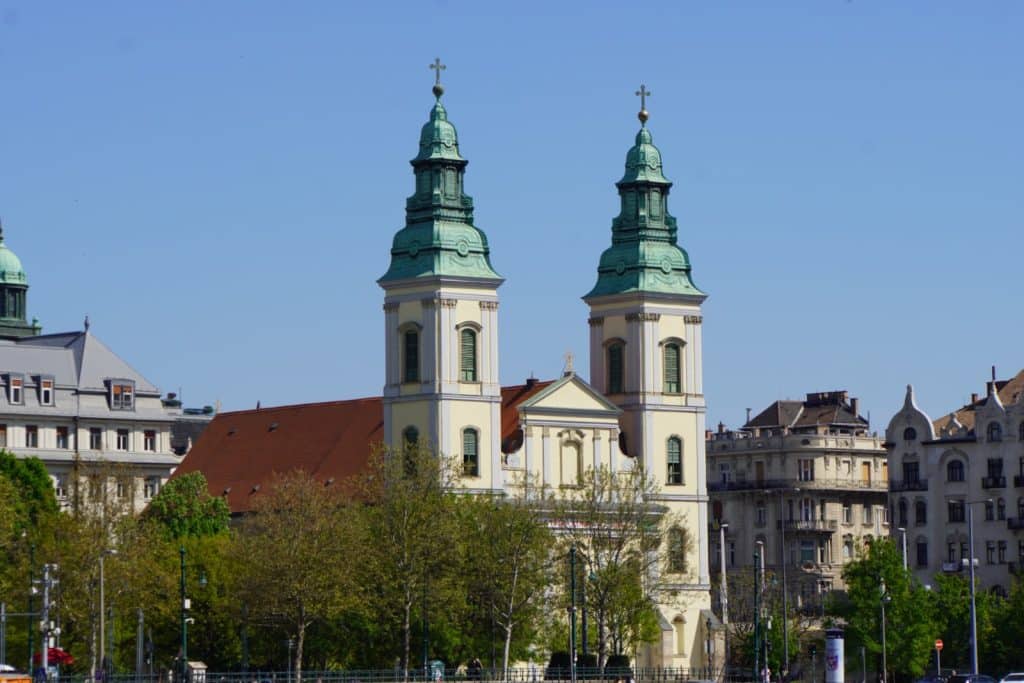 Die Innerstädtische Pfarrkirche in Budapest mit ihren zwei markanten Türmen mit grünen Kuppeln und dem roten Ziegeldach steht vor einem klaren blauen Himmel, umgeben von Bäumen und historischen Gebäuden.