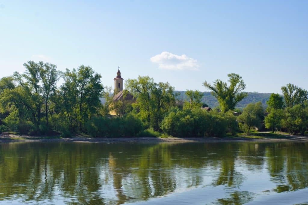 Blick von einem Schiff während einer Flusskreuzfahrt auf eine am Ufer gelegene Kirche mit rötlichem Turm, die von grünen Bäumen umgeben ist und sich im Wasser spiegelt.