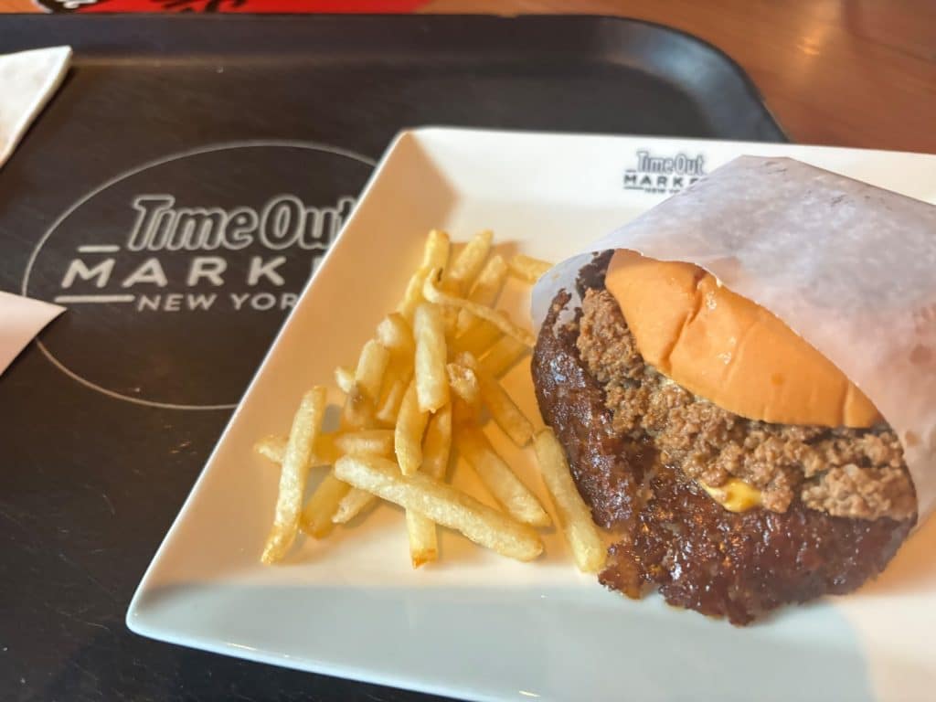 Ein Burger mit knusprig gebratenem Fleisch und eine Portion Pommes frites werden auf einem weißen Teller im Time Out Market New York serviert.
