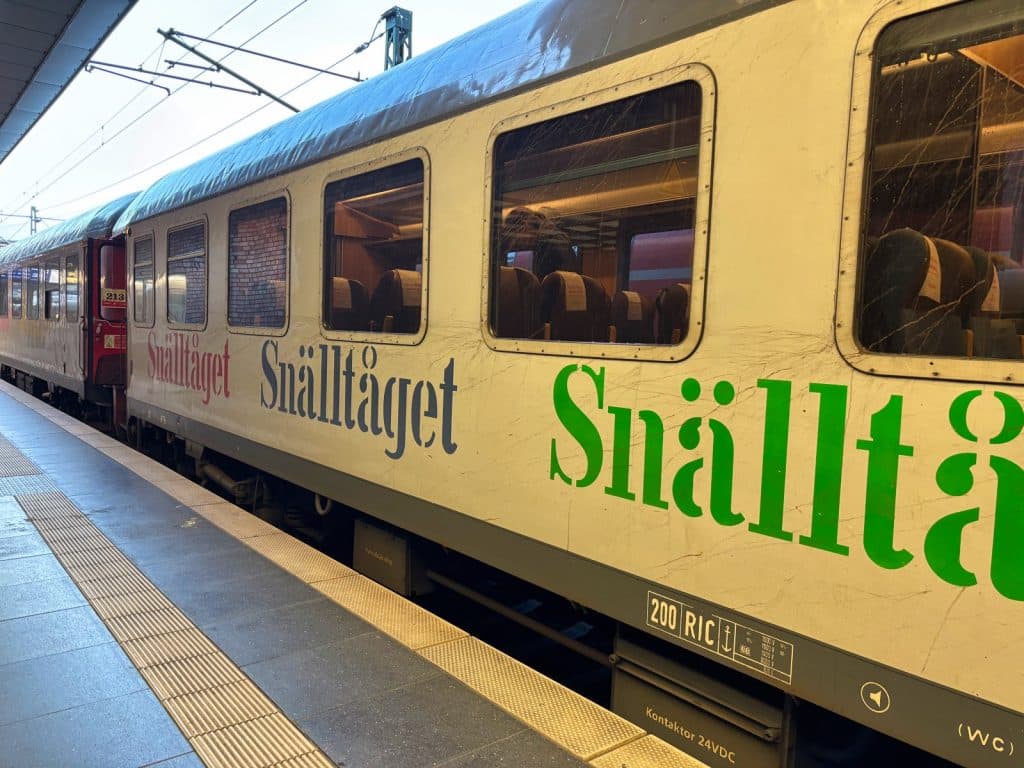 Ein weißer Snälltåget-Reisezug mit großen grünen und blauen Schriftzügen steht an einem Bahnsteig in Berlin. Die Nahaufnahme zeigt die Waggons mit Fenstern, durch die man die Sitzreihen im Inneren erkennen kann.