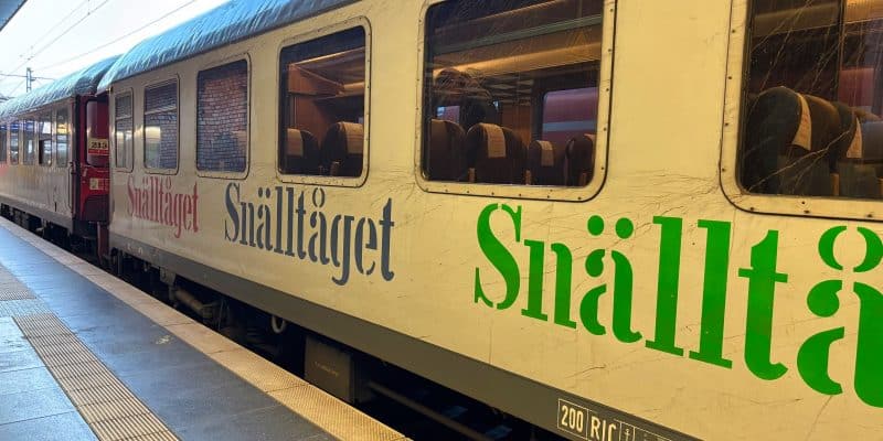 Ein weißer Snälltåget-Reisezug mit großen grünen und blauen Schriftzügen steht an einem Bahnsteig in Berlin. Die Nahaufnahme zeigt die Waggons mit Fenstern, durch die man die Sitzreihen im Inneren erkennen kann.