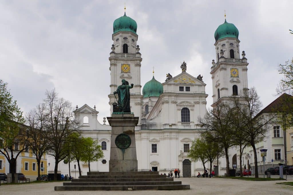 Außenansicht des Passauer Doms St. Stephan mit seinen zwei markanten weißen Türmen mit grünen Zwiebelkuppeln, davor ein großer Platz mit einem zentralen Denkmal auf einem Steinsockel.
