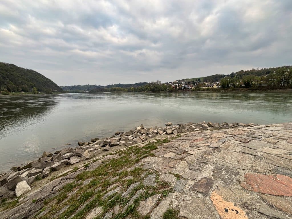 Blick vom gepflasterten Ufer auf das Dreiflüsseeck in Passau, wo Donau, Inn und Ilz unter einem bewölkten Himmel zusammenfließen.