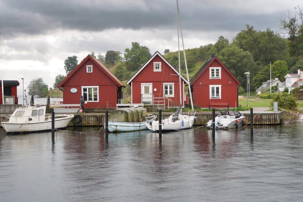 Drei traditionelle rote Holzhäuser mit weißen Fensterrahmen stehen am Hafenbecken der Insel Ven bei Landskrona, während davor verschiedene Segel- und Motorboote an einem Steg im ruhigen Wasser liegen.